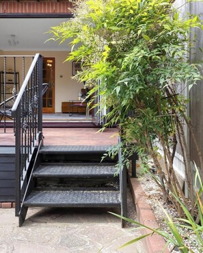 Un escalier menant à une terrasse en bois, agrémentée d’une rambarde en métal, le tout entouré de verdure et avec un bâtiment visible au loin.