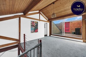 A spacious room with a high ceiling, wooden beams, a large window, and a red door.