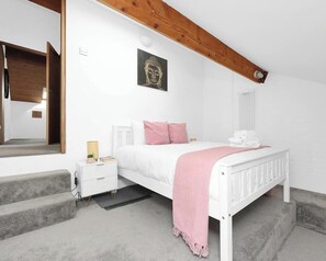 A bedroom with a white bed, a nightstand, a window, a wooden ceiling beam, and a Buddha artwork on the wall.