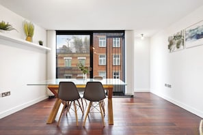 Un área de comedor moderna con una mesa de vidrio, dos sillas y vistas a un edificio de ladrillo a través de una puerta corredera.