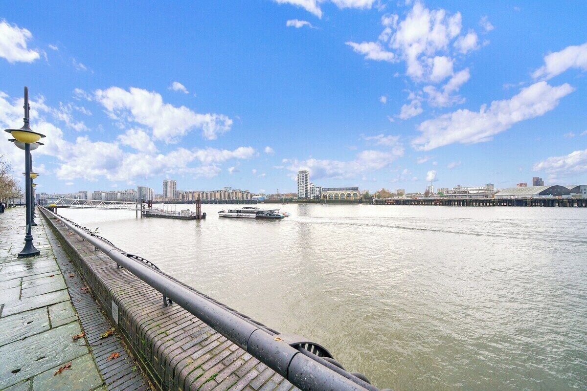 A riverside walkway with a metal railing, a boat on the water, and a cityscape in the background.