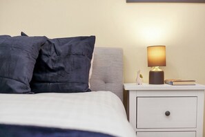A neatly made bed with dark bedding, a bedside lamp, a small decorative item, and a book on a white nightstand.