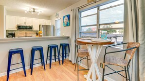A kitchen with white cabinets, a microwave, and a refrigerator. There are four blue stools and a wicker chair around a small table. A sign on the wall reads "Beach House". - 1 Mi to Boardwalk & Dtwn: Myrtle Beach Condo (Myrtle Beach)