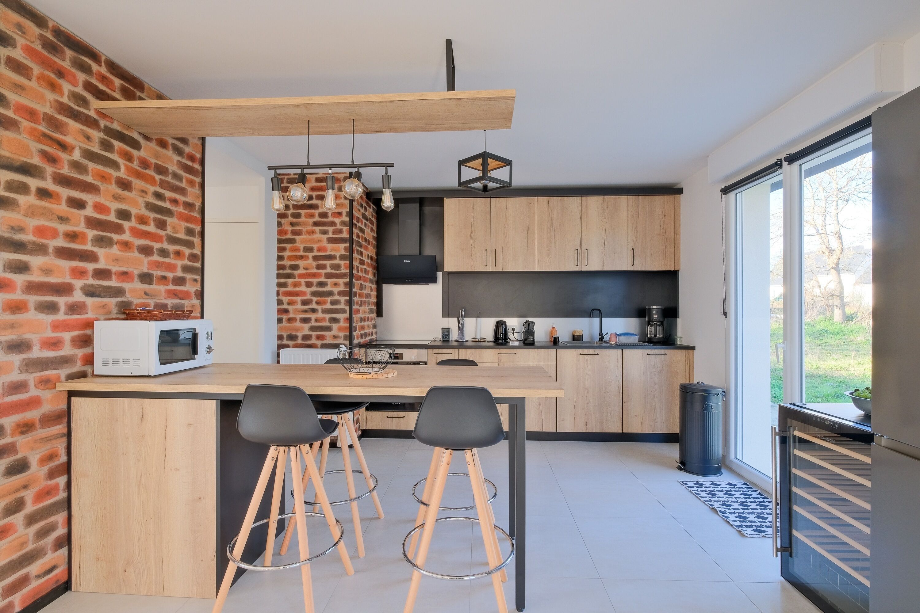 A modern kitchen with a brick wall, wooden cabinets, a large island with stools, and a view through a sliding door.