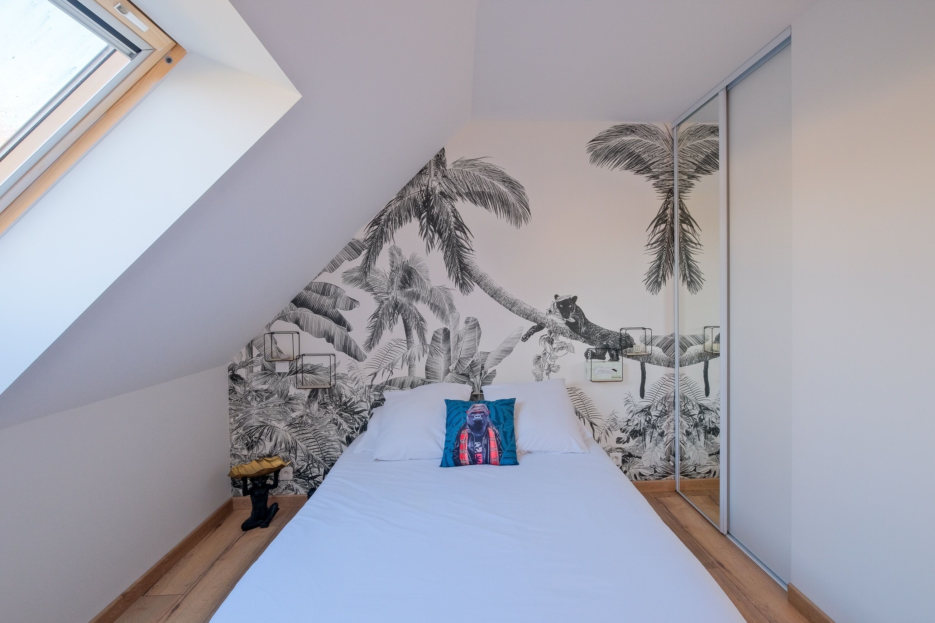 A bedroom with a sloped ceiling, a bed with a blue pillow, a nightstand, a small table with a lamp, and a wall mural of palm trees.