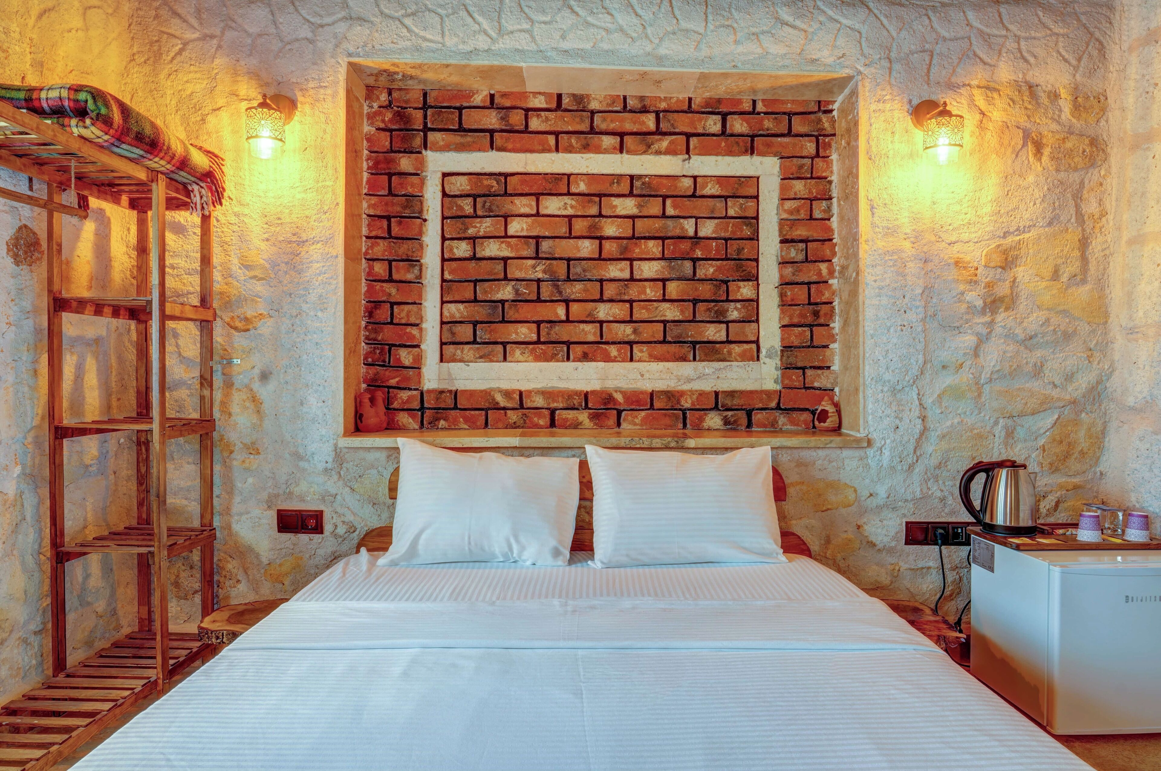 Une chambre avec un mur au motif de briques, un lit recouvert de draps blancs, une échelle en bois, une étagère avec une couverture et une petite table sur laquelle se trouvent une bouilloire et des tasses.