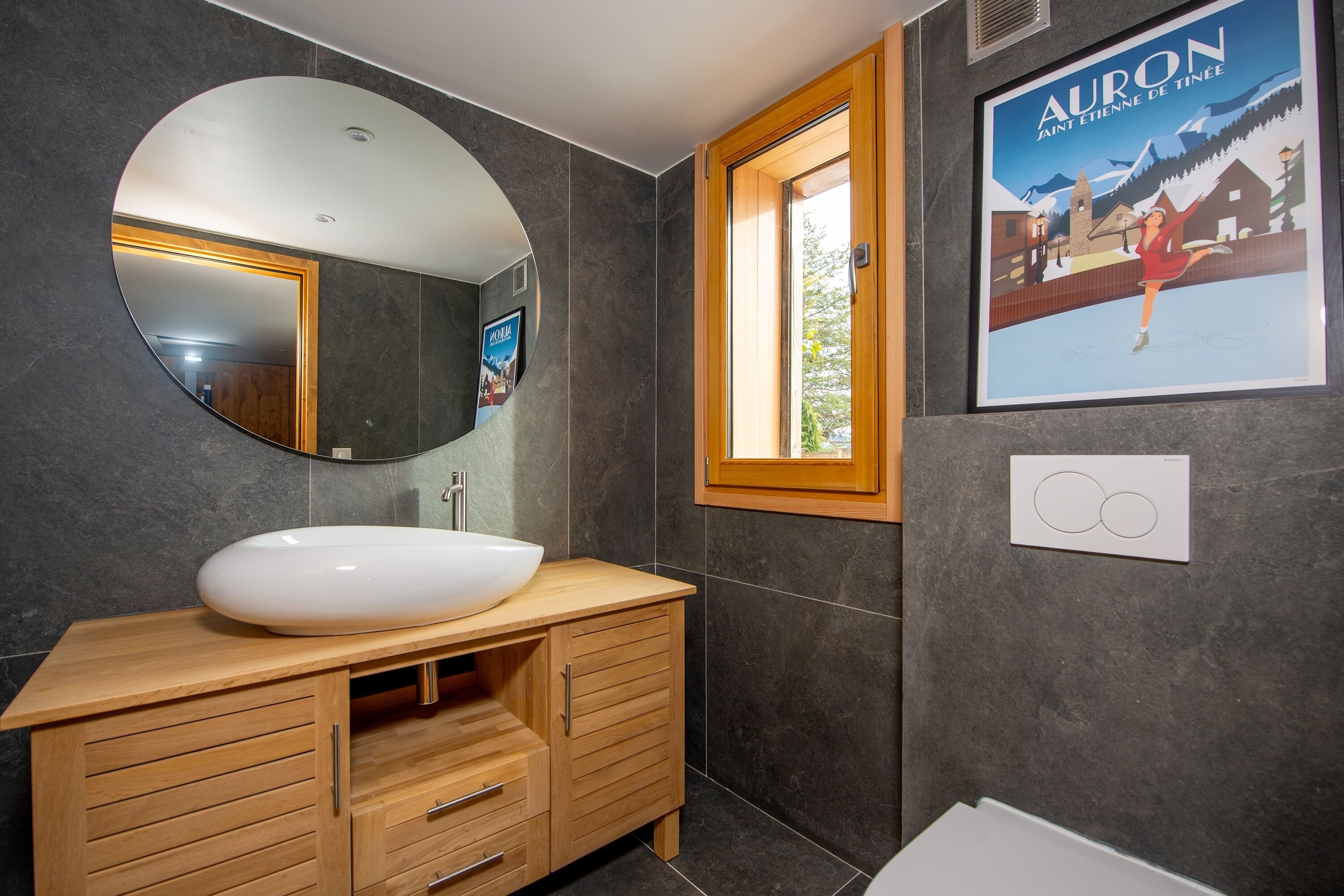 A modern bathroom with a large oval sink, wooden vanity, a round mirror, a window with a wooden frame, and a poster on the wall.