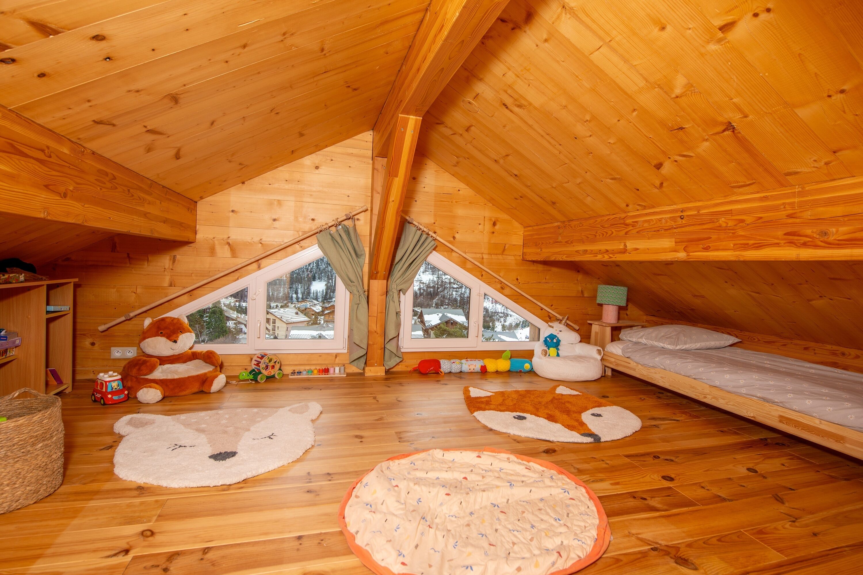 A cozy attic bedroom with wooden walls and a sloped ceiling, featuring a bunk bed, a window with a view of snow-covered trees, and various children's toys and rugs.