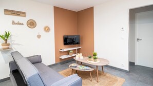 Ein modernes Wohnzimmer mit grauem Sofa, einem runden Holztisch, einem flachbildfernseher an der Wand und einem kleinen Beistelltisch mit Dekoration.