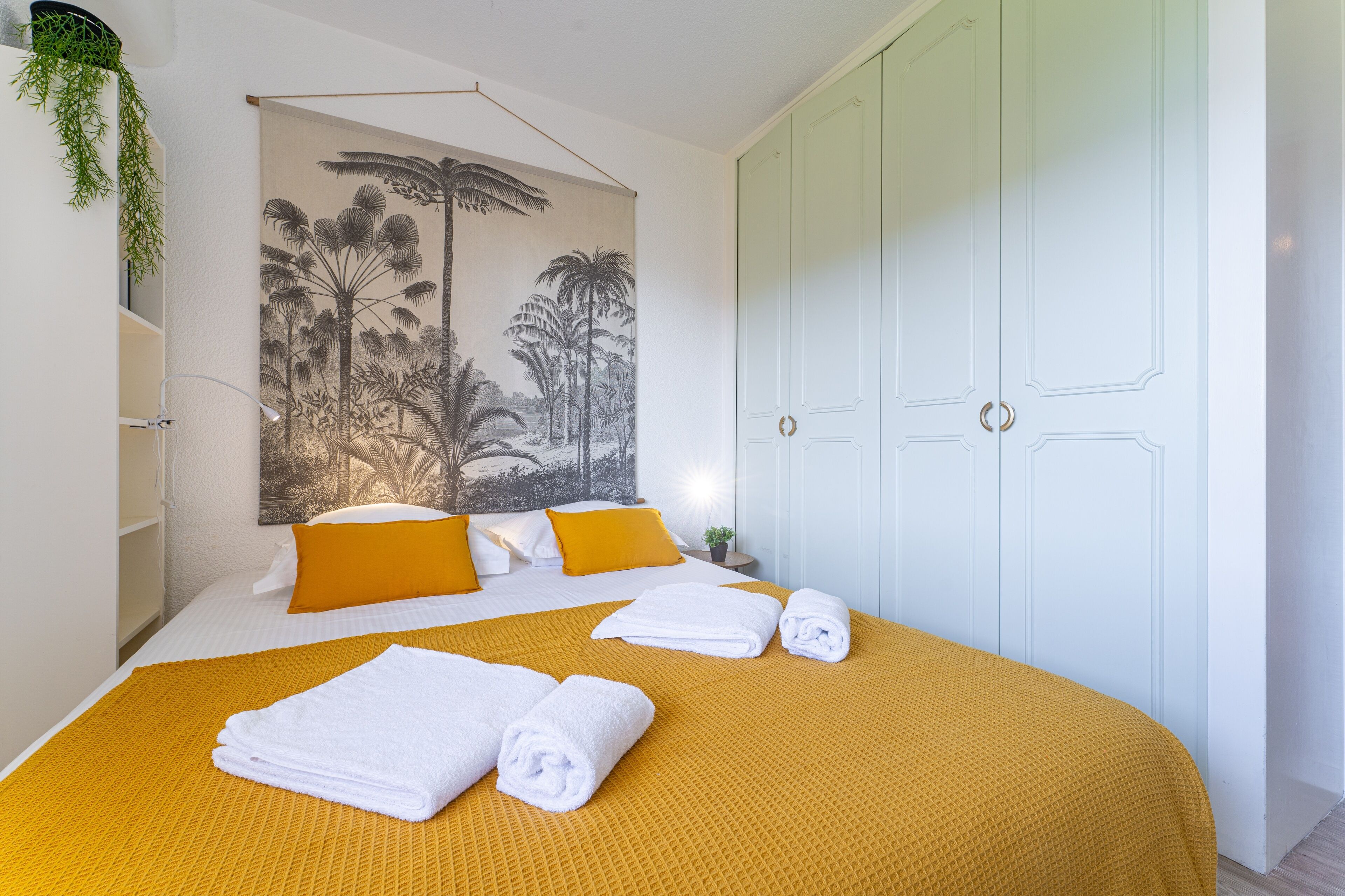 Habitación de hotel con cama, paredes blancas, un cuadro grande de palmeras, armario blanco y planta colgante.