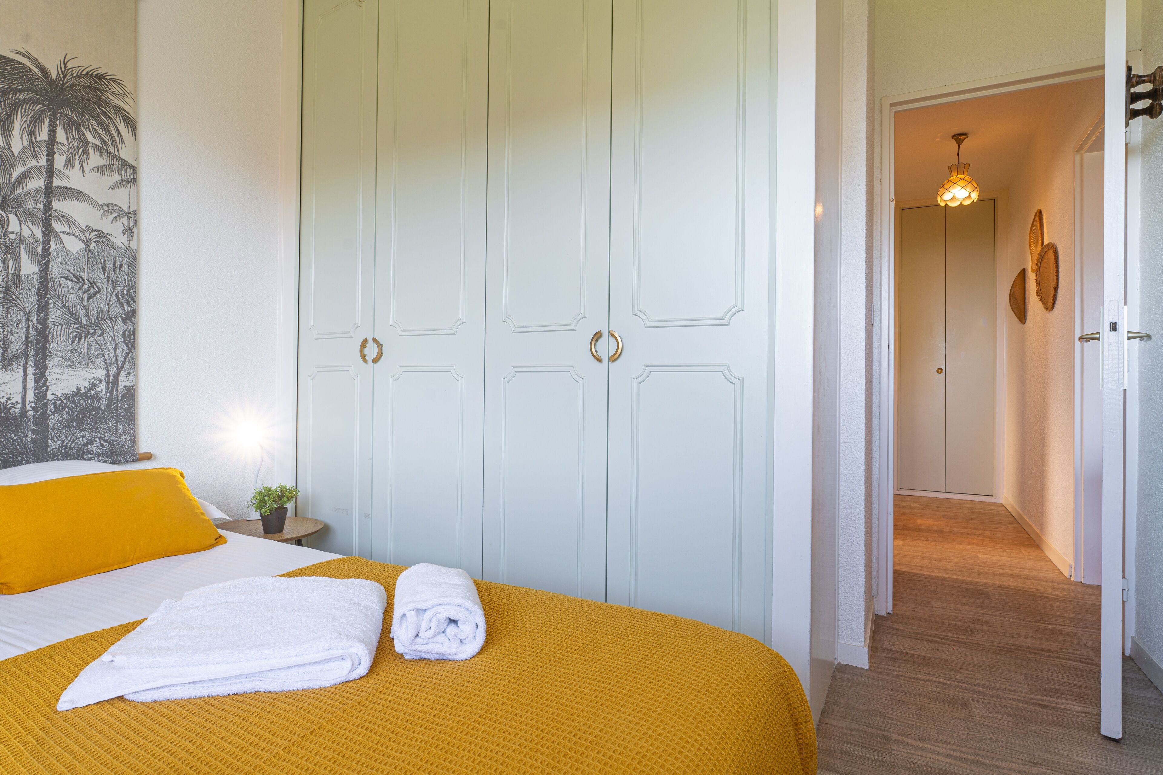 Hotellihuone, jossa on sänky, valkoinen ovi, käytävä ja valaisin.