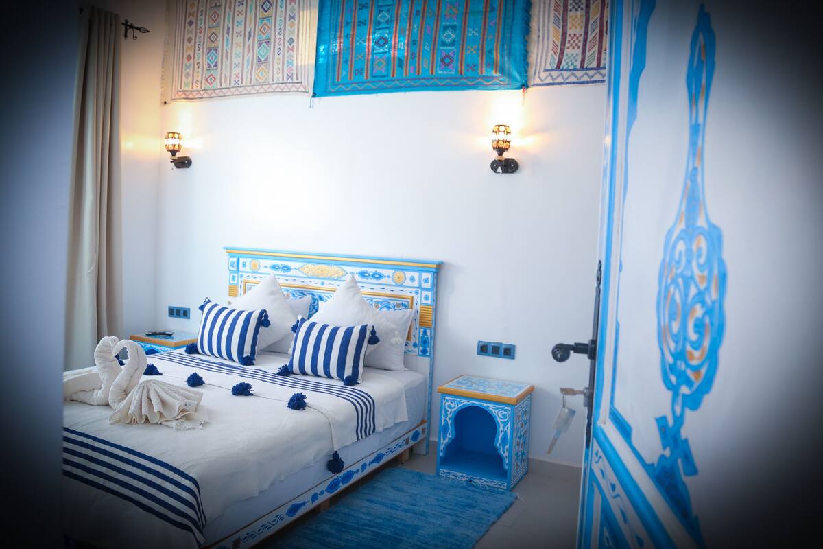 A bedroom with a bed, pillows, a nightstand, a wall-mounted lamp, and a decorative wall hanging.