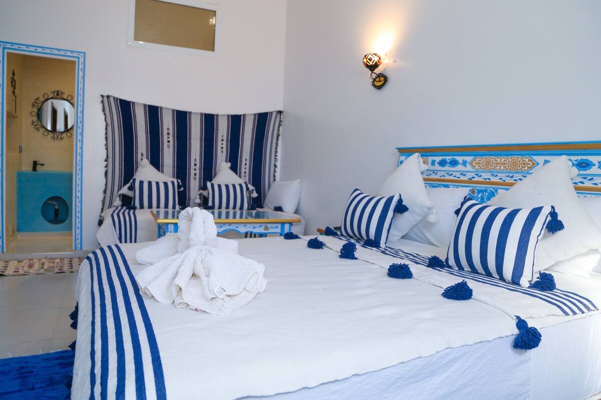 A hotel room with a bed, pillows, a bedside table, a mirror, and a wall-mounted light fixture.