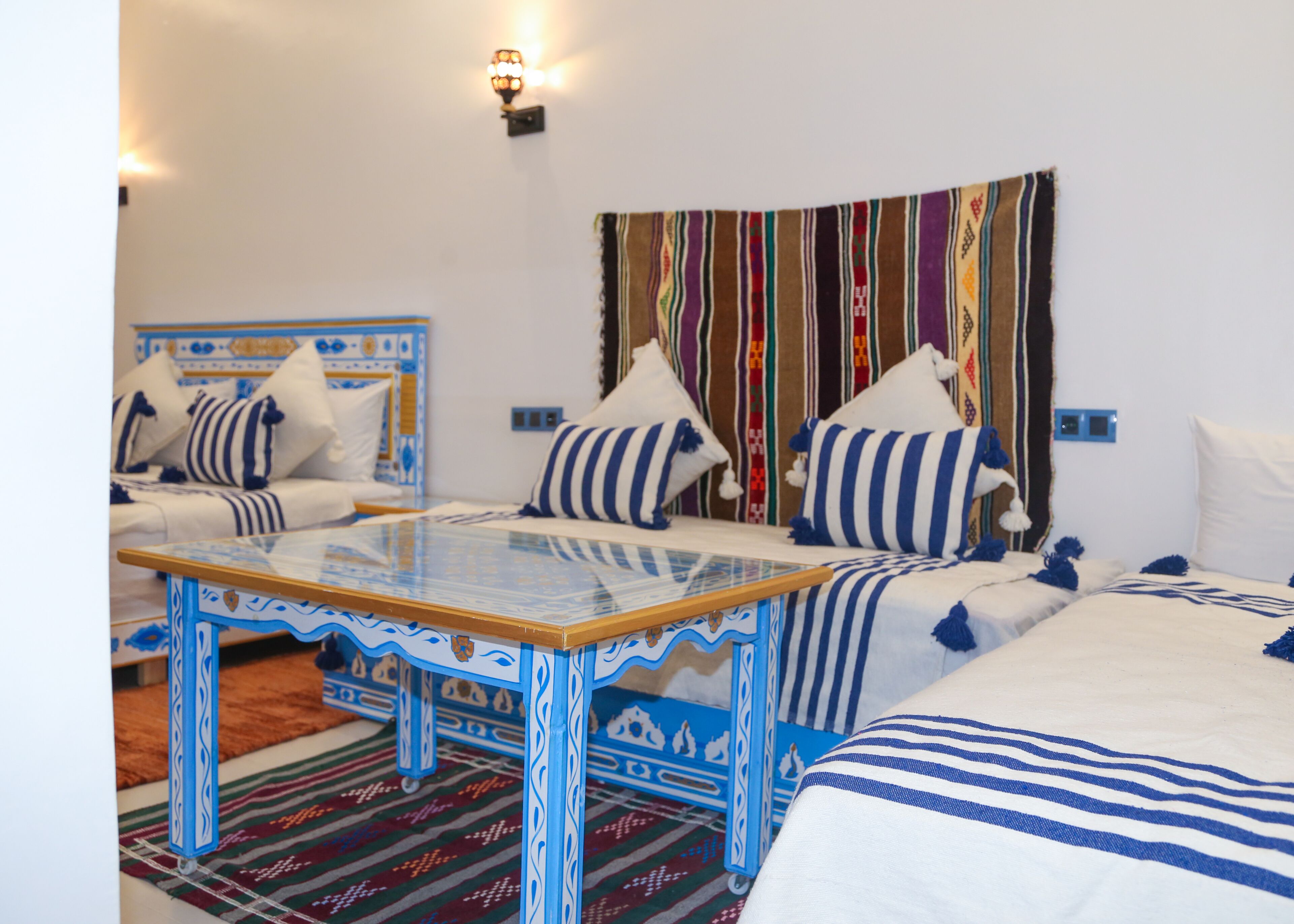A room with two beds, a glass-top table, and a colorful striped tapestry on the wall.
