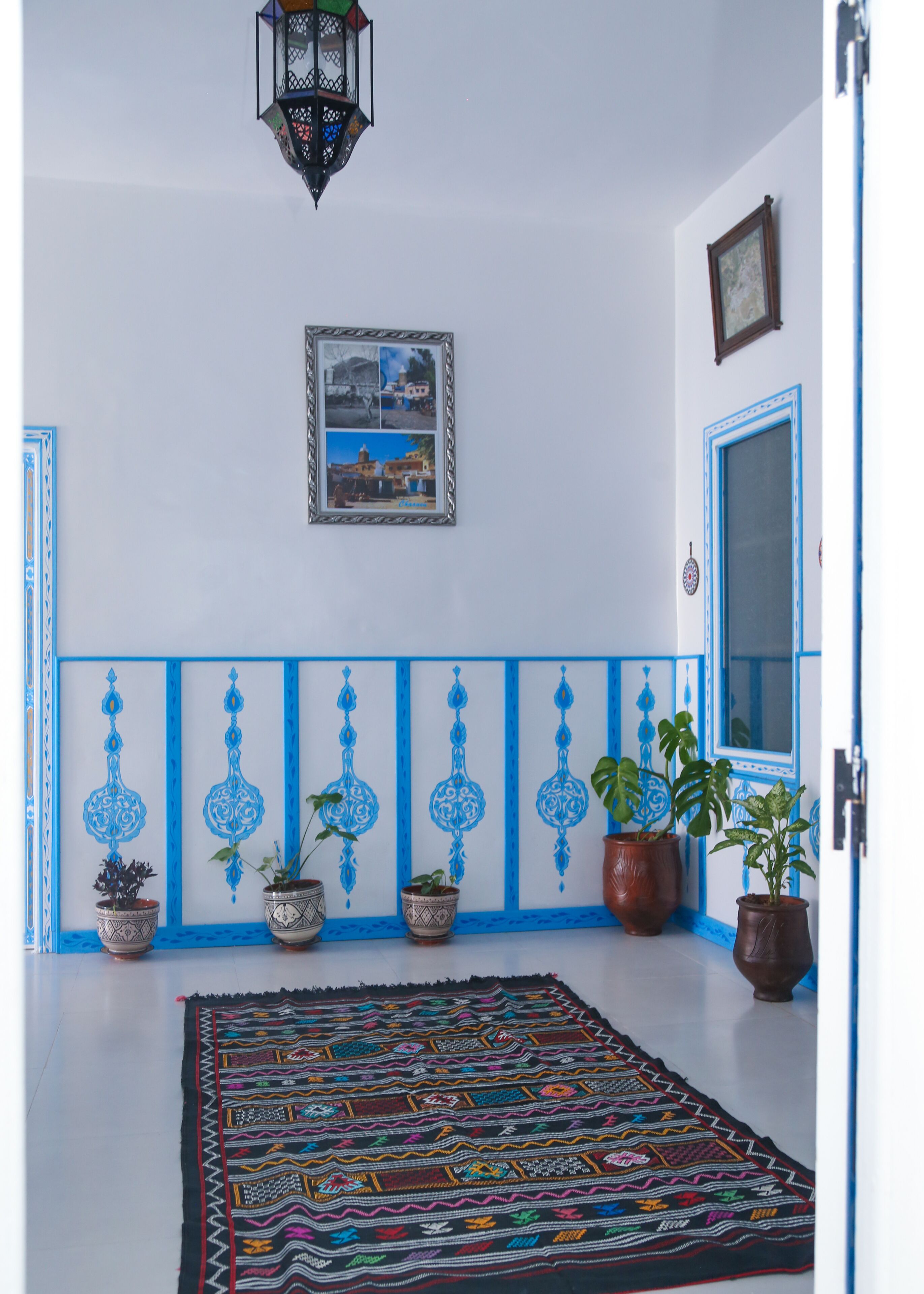 A hallway with a patterned rug, potted plants, a framed picture on the wall, and a door with blue trim.