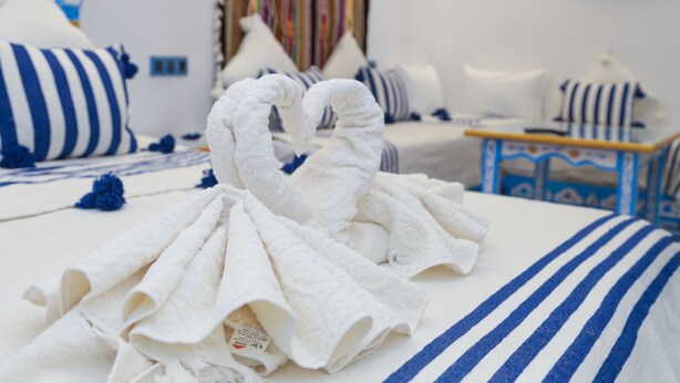 A hotel room with a bed featuring blue and white striped bedding, a white swan-shaped towel, and a colorful striped curtain. - Casa Blue Pearl (Chefchaouen)