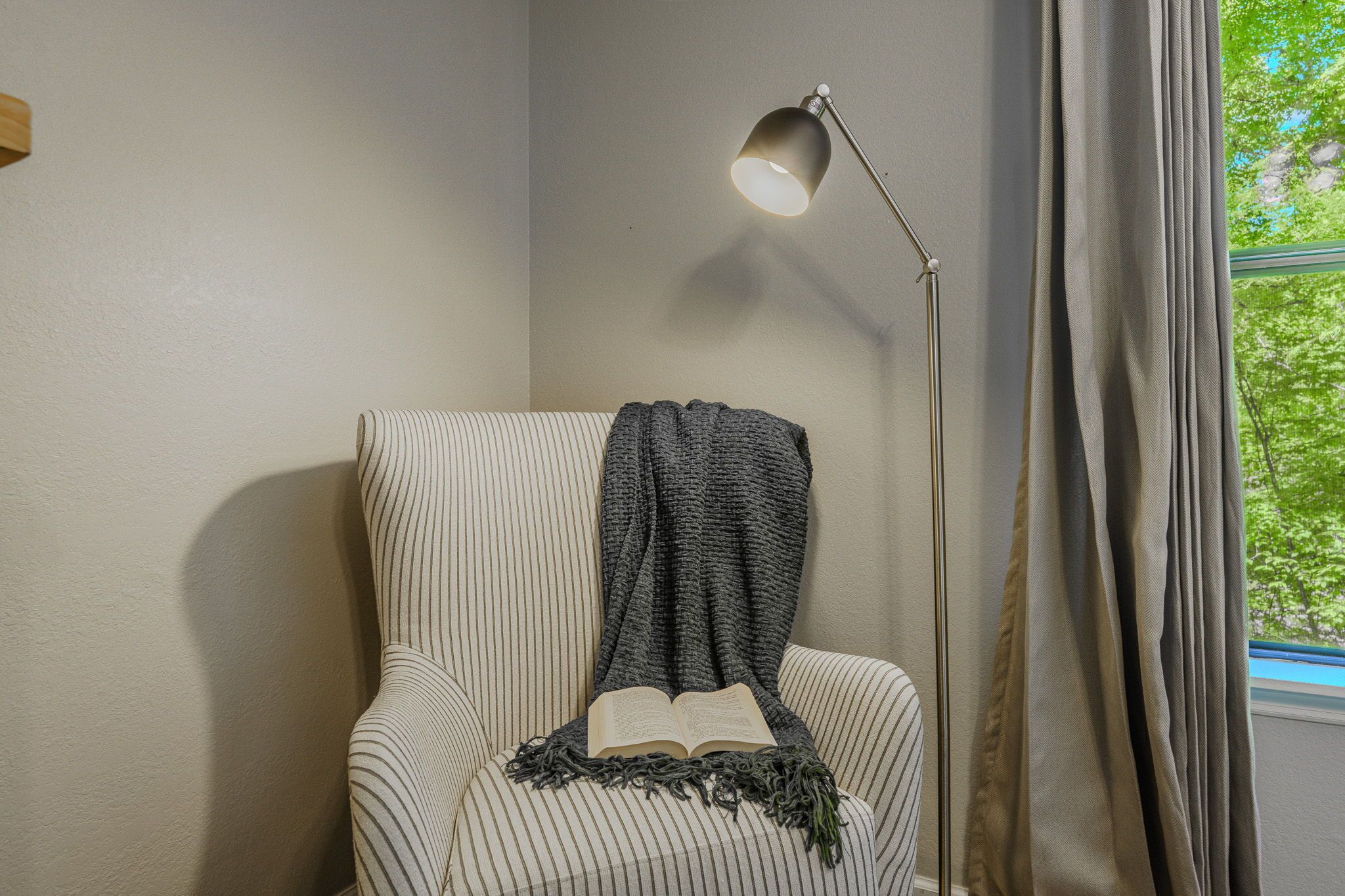A striped armchair with a book and a throw blanket, a floor lamp, and a window with curtains.
