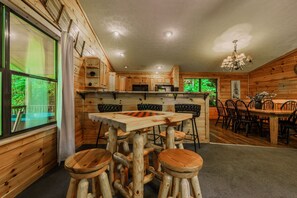 A rustic cabin interior with a wooden dining table, bar stools, and a kitchen area with wooden cabinets.