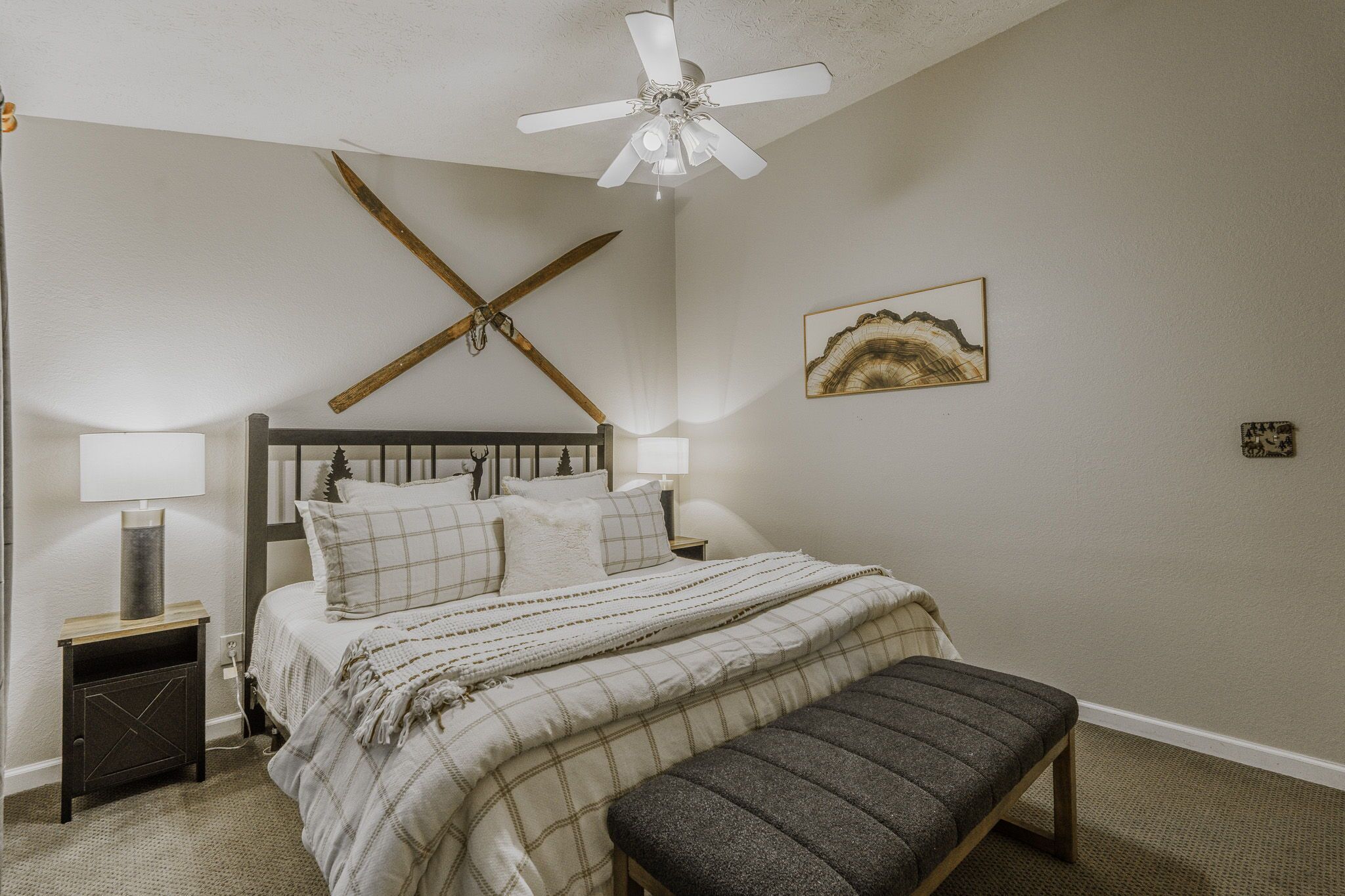 A bedroom with a bed, bedside lamps, a heater, a bench, a ceiling fan, and a wall-mounted artwork.