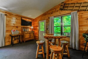 A wooden interior with a dining table, stools, a television, and a window with a view of greenery.