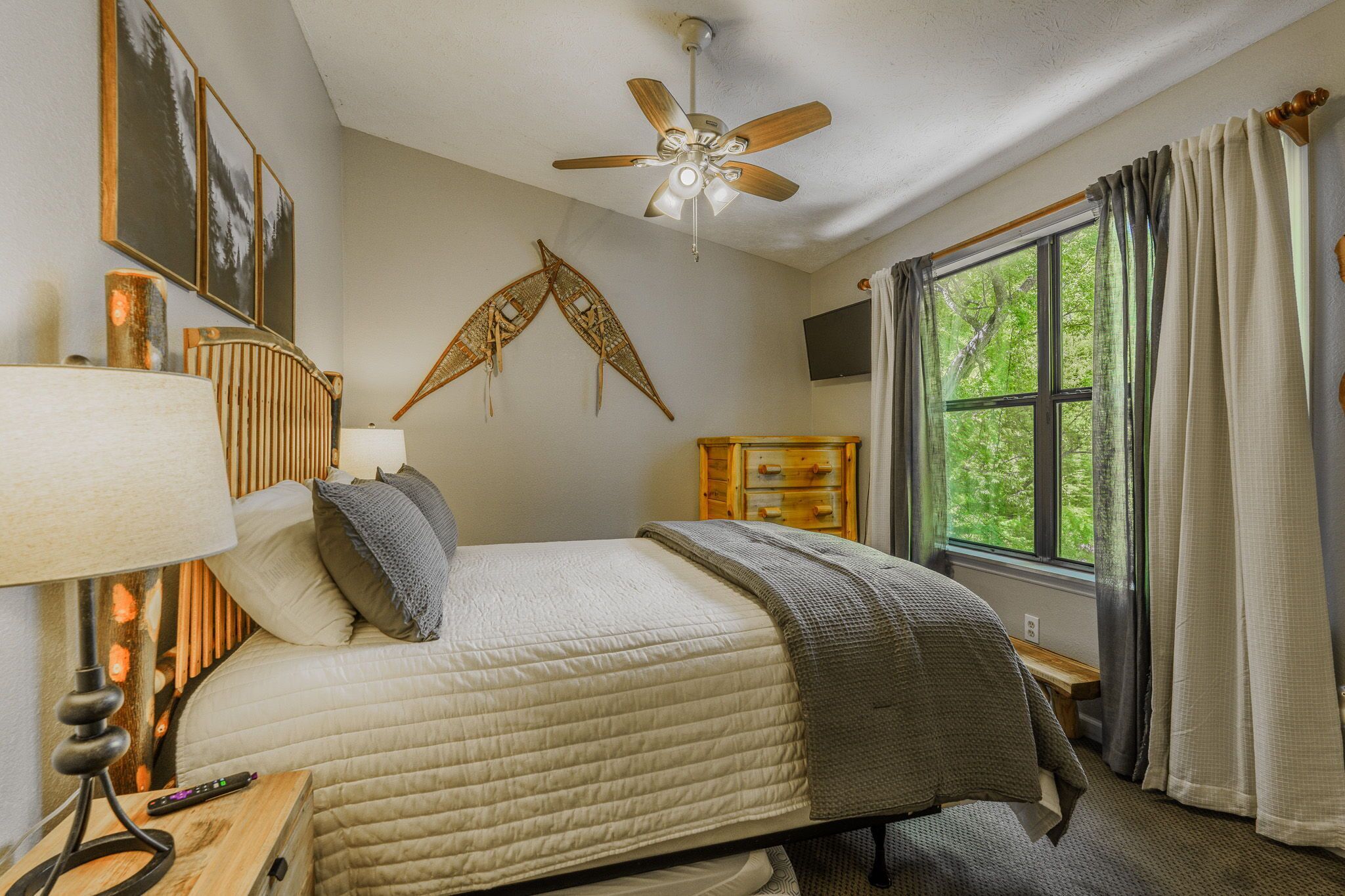 A bedroom with a bed, a ceiling fan, a window with curtains, a wooden dresser, and wall-mounted artwork.
