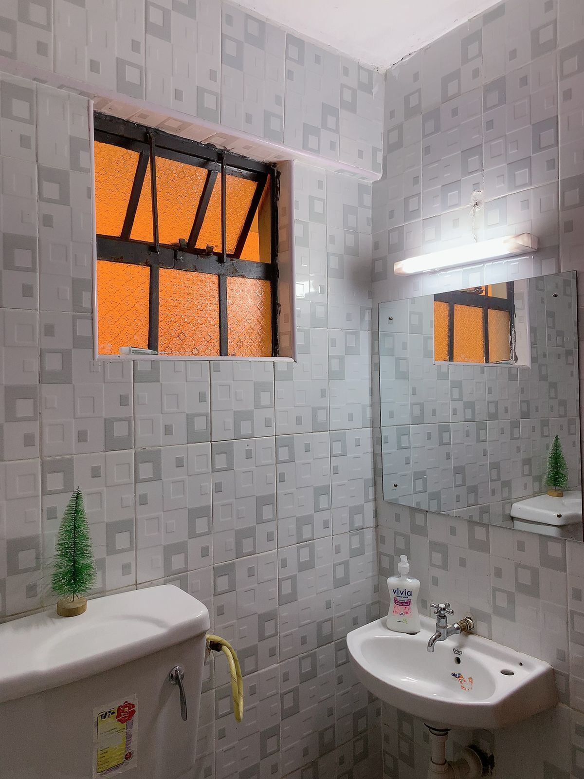 Une salle de bain avec des carreaux décorés, une fenêtre à cadre noir, un petit sapin de Noël, des toilettes, un lavabo et un miroir.