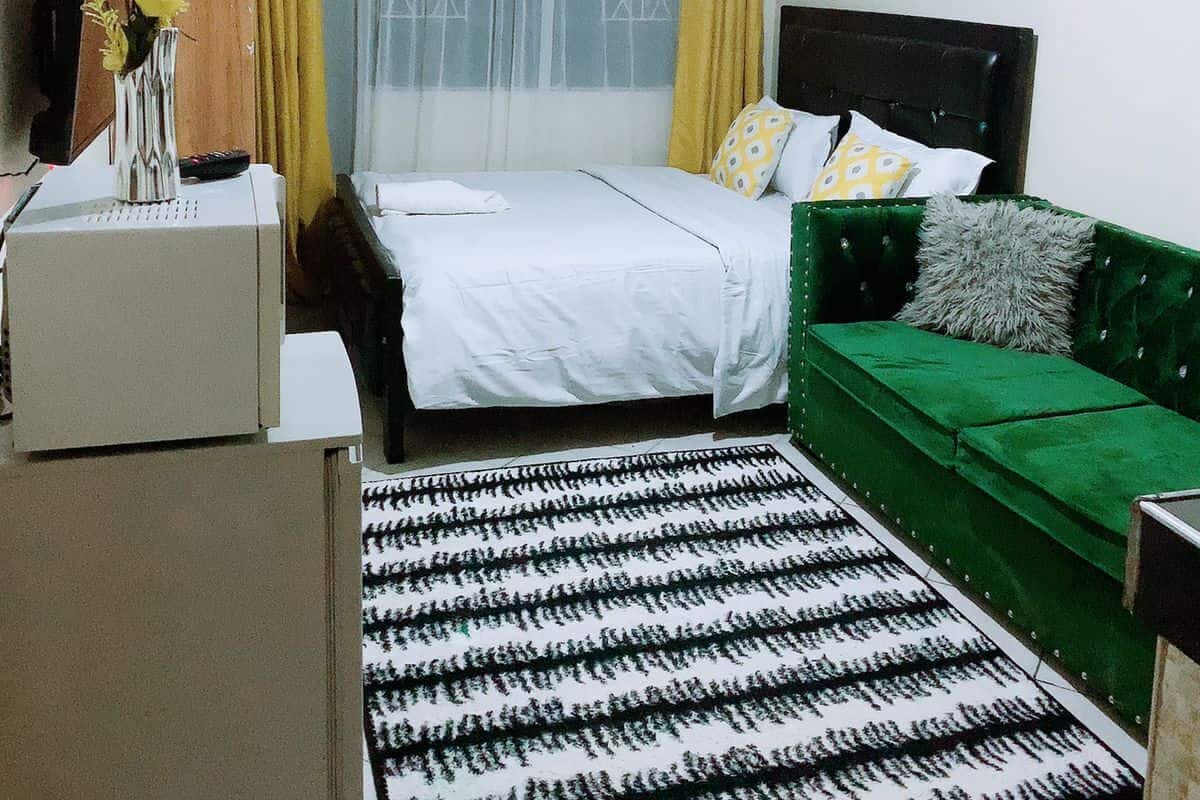 A bedroom with a bed, a green sofa, a small table with a vase, and a patterned rug.
