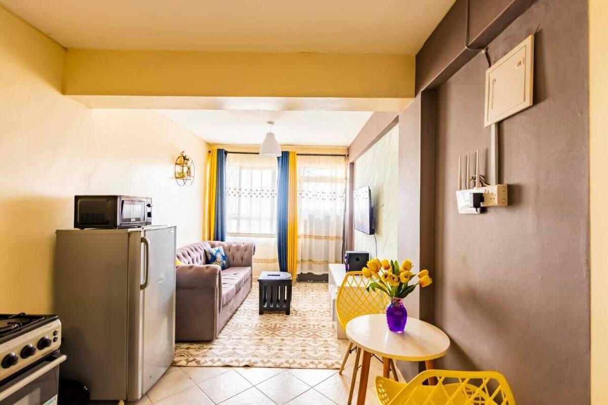 A compact living space with a kitchenette, a sofa, a dining table with yellow chairs, and a TV mounted on the wall.