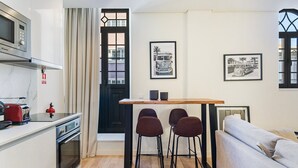 A compact kitchen with a microwave, toaster, and oven. There is a dining area with a table and four chairs. The room has a black door with a window, and two framed pictures on the wall. - GuestReady - Secret in Porto (Porto)