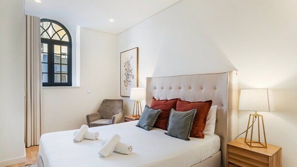 A neatly made bed with pillows, a chair, bedside lamps, and a window with curtains. - GuestReady - Secret in Porto (Porto)