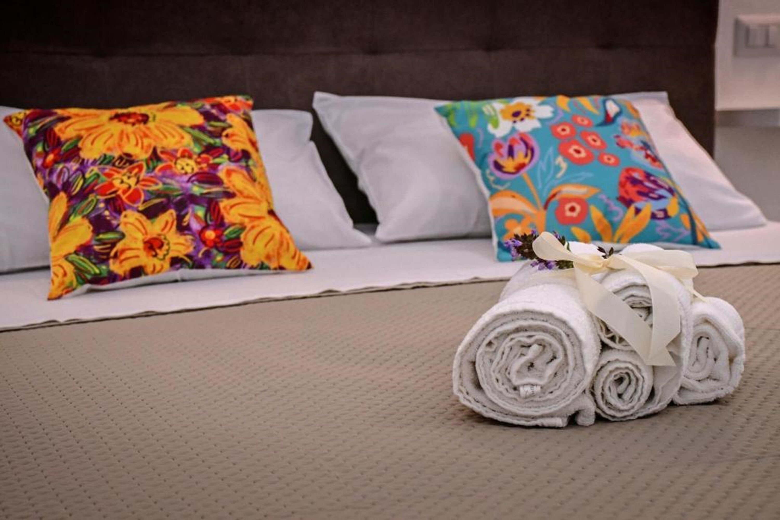 Una cama con almohadones florales de colores y una toalla blanca enrollada sobre una colcha beige con textura.