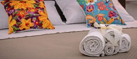 Una cama con almohadones florales de colores y una toalla blanca enrollada sobre una colcha beige con textura.