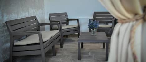 Una habitación con cuatro sillas de color oscuro y una mesita, un jarrón con flores azules, y una cortina recogida lateralmente.