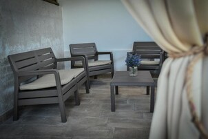 Una habitación con cuatro sillas de color oscuro y una mesita, un jarrón con flores azules, y una cortina recogida lateralmente.