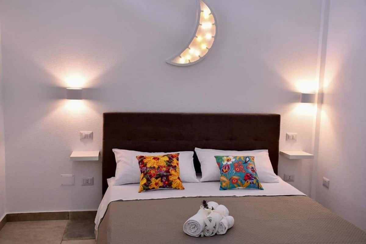 A neatly made bed with two colorful pillows, a moon-shaped light fixture above, and two wall-mounted lights on either side.