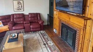 A living room with a red leather sofa, a fireplace, a television mounted on a wooden wall, and a ceiling fan. - Retro Ranchhouse (Sanford)
