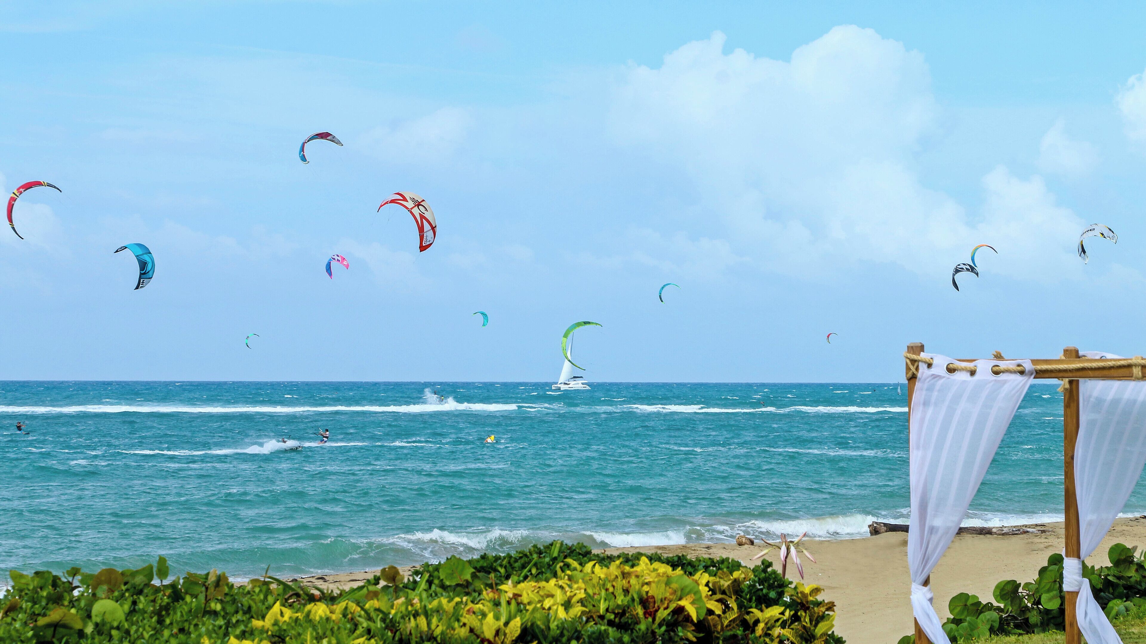 Ein Strand mit einem Parasailing-Boot in der Ferne und mehreren Drachen am Himmel.