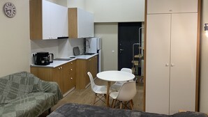 Una cocina compacta con armarios blancos, una cafetera y un lavabo. Hay un comedor con una mesa y sillas blancas, y un sofá en primer plano.