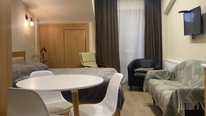 Una habitación compacta con cama, una mesa de comedor con sillas, un sofá, un televisor fijado a la pared y una ventana con cortinas.