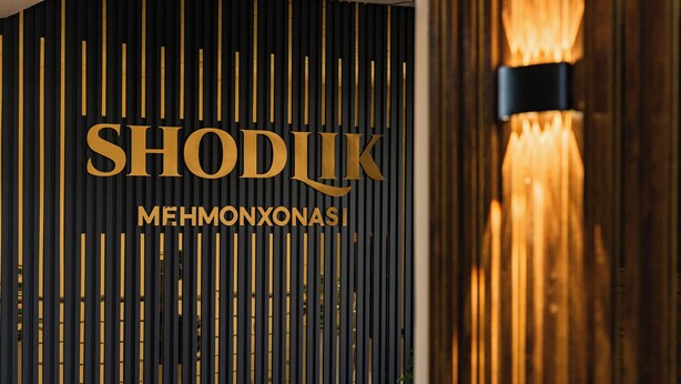 A wall with vertical wooden slats and the text "SHODUK MEHMONXONASI" in gold letters. - Hotel Shodlik  (Samarkand)