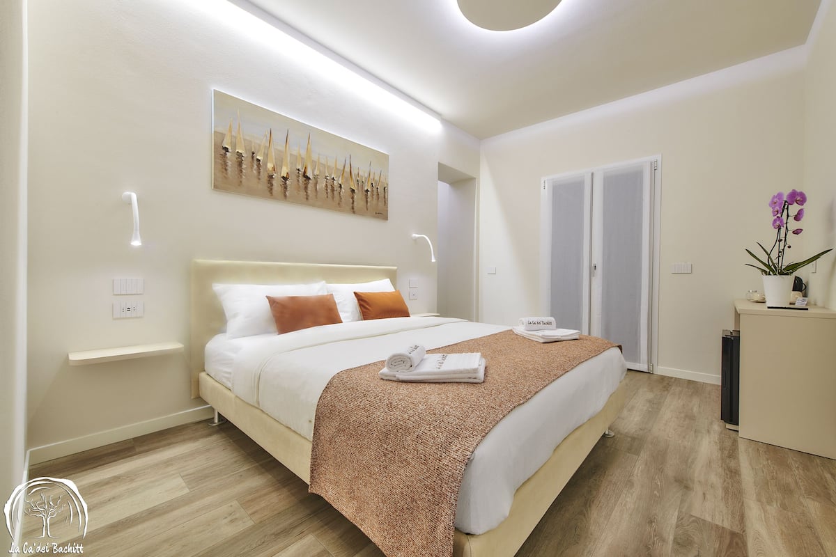 A modern bedroom with a large bed, a bedside table, a nightstand, a wall-mounted painting, a door, and a potted plant.