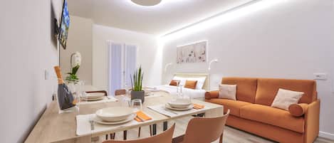 A modern living room with a sofa, dining table, and a flat-screen TV mounted on the wall.