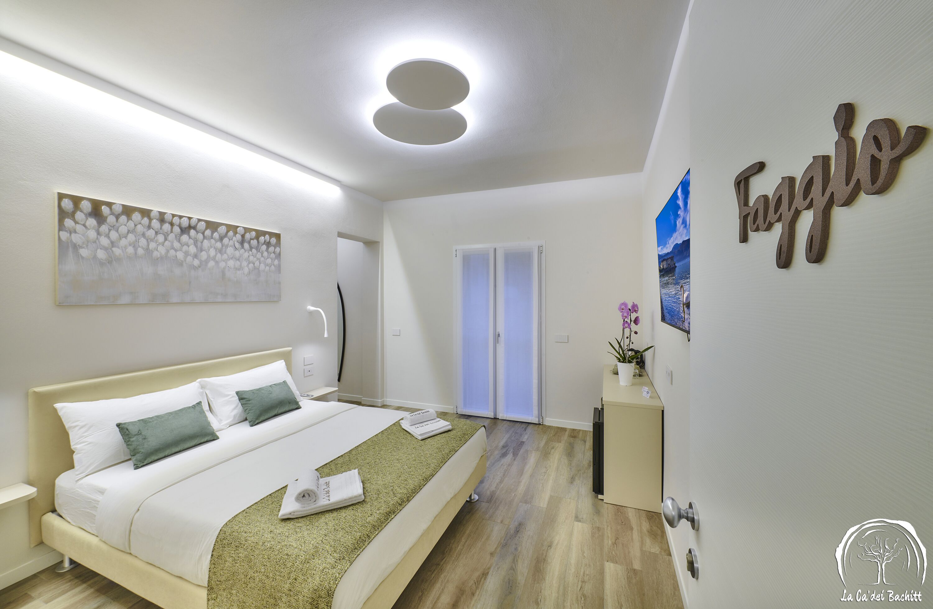 Una habitación de hotel moderna con una cama bien hecha, una obra de arte en la pared, una puerta con paneles de vidrio esmerilado y una placa identificativa en la pared.