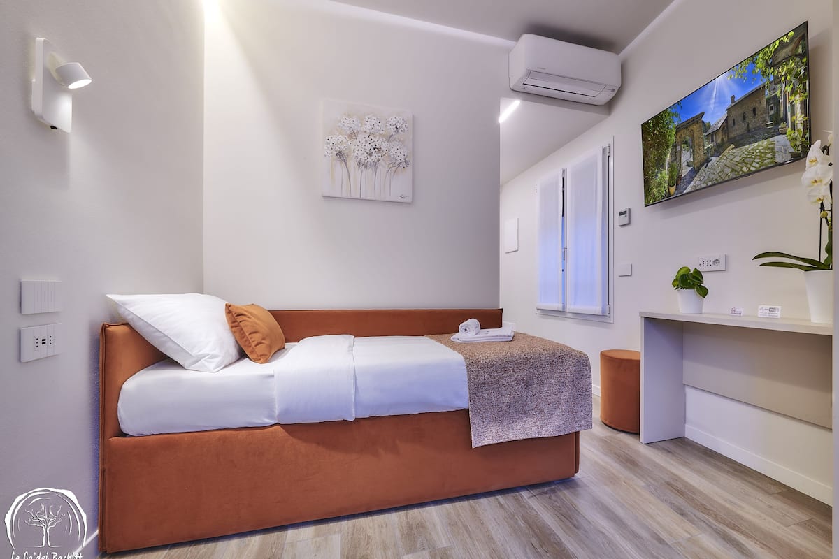 A modern bedroom with a brown sofa bed, white bedding, a wall-mounted air conditioner, a flat-screen TV, and a small potted plant.