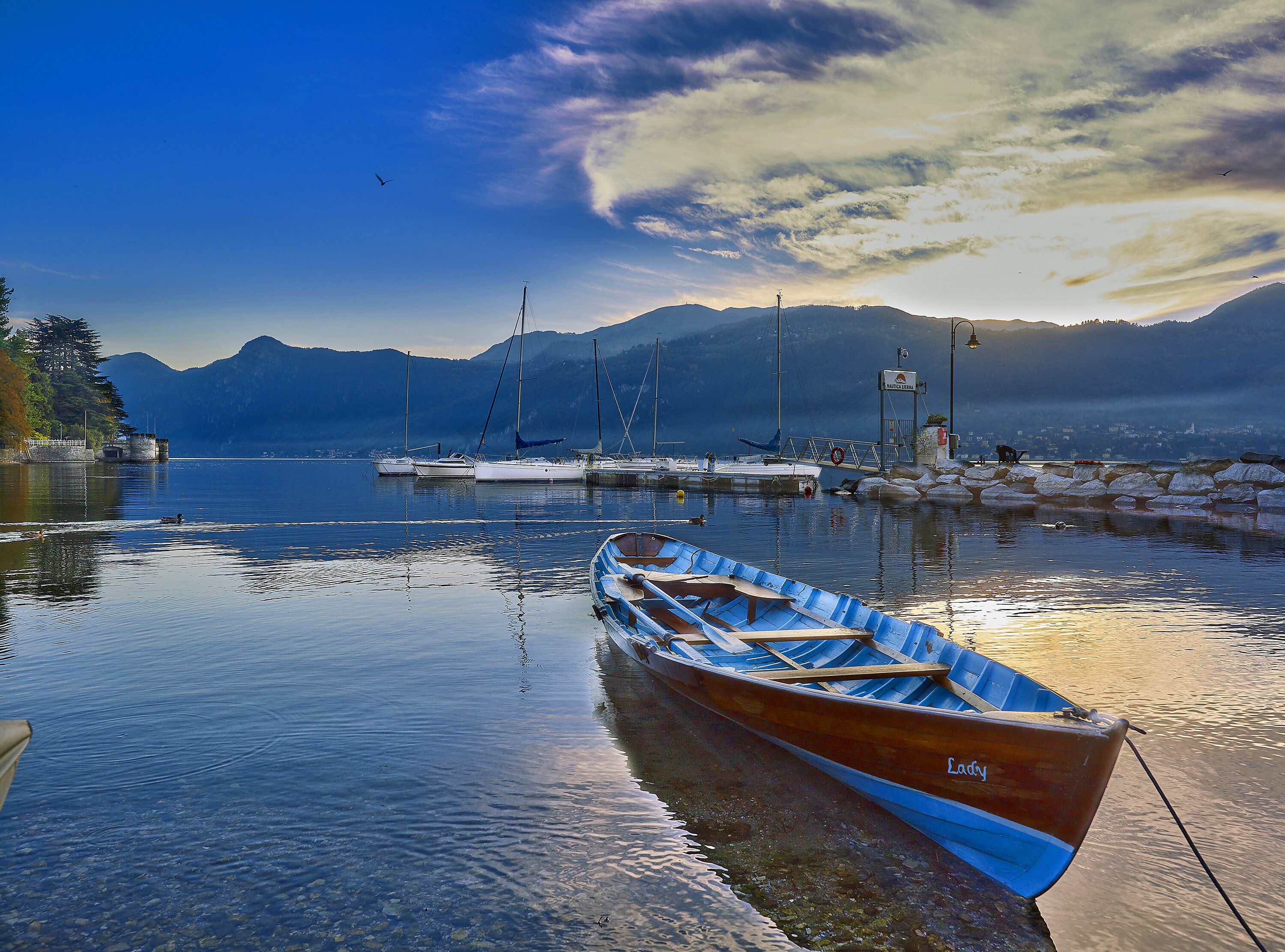 Un lago tranquilo con barcos, un muelle y montañas al fondo.