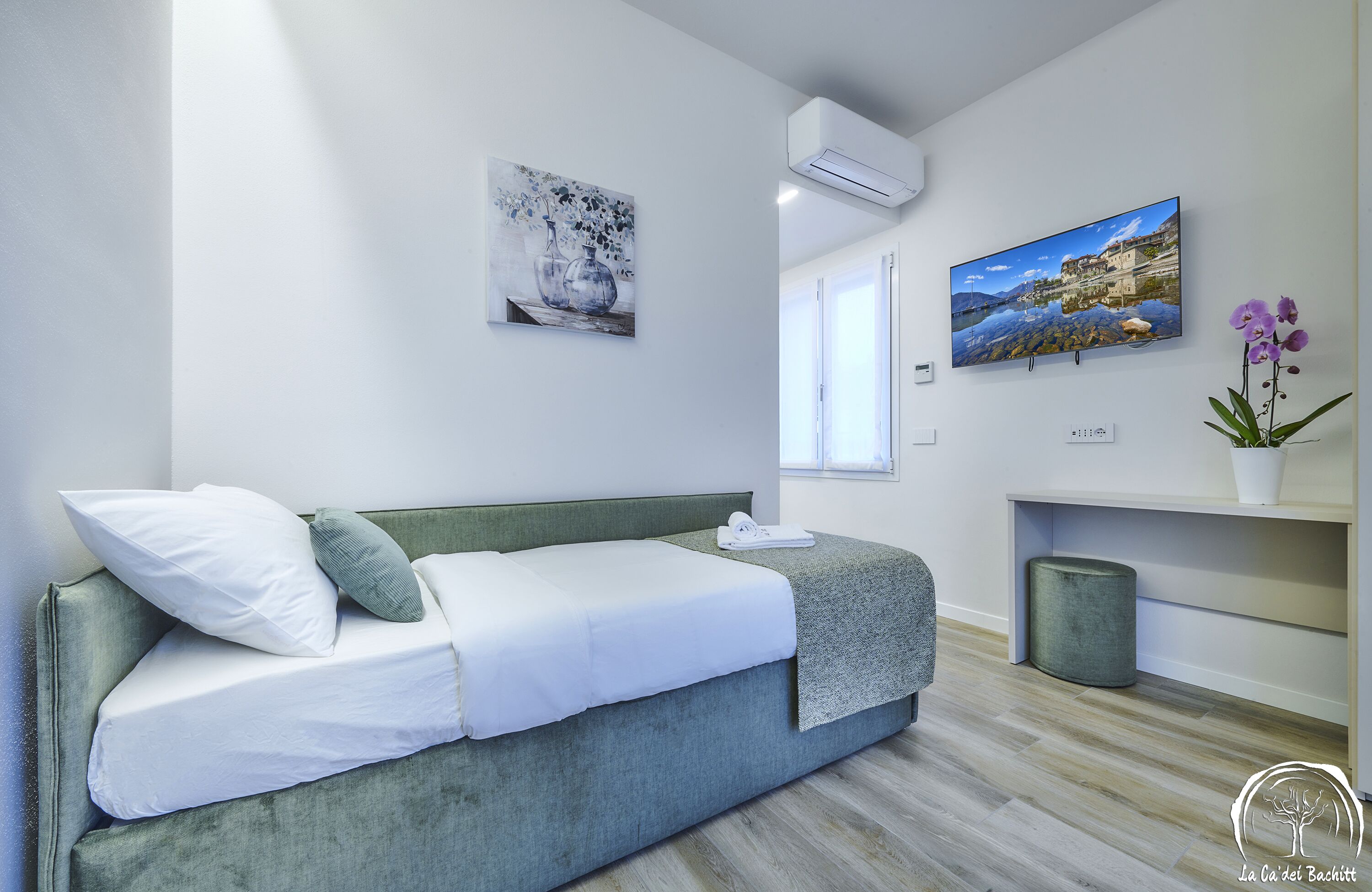 A modern bedroom with a green sofa bed, white bedding, a mounted TV, and a small potted orchid.