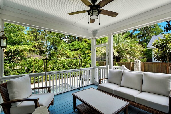 A covered porch with a ceiling fan, white outdoor furniture, and a view of a lush garden. - Beautiful 4 bed, 3.5 bath home w/pool and hot tub. Walk to Village area! (Saint Simons Island)