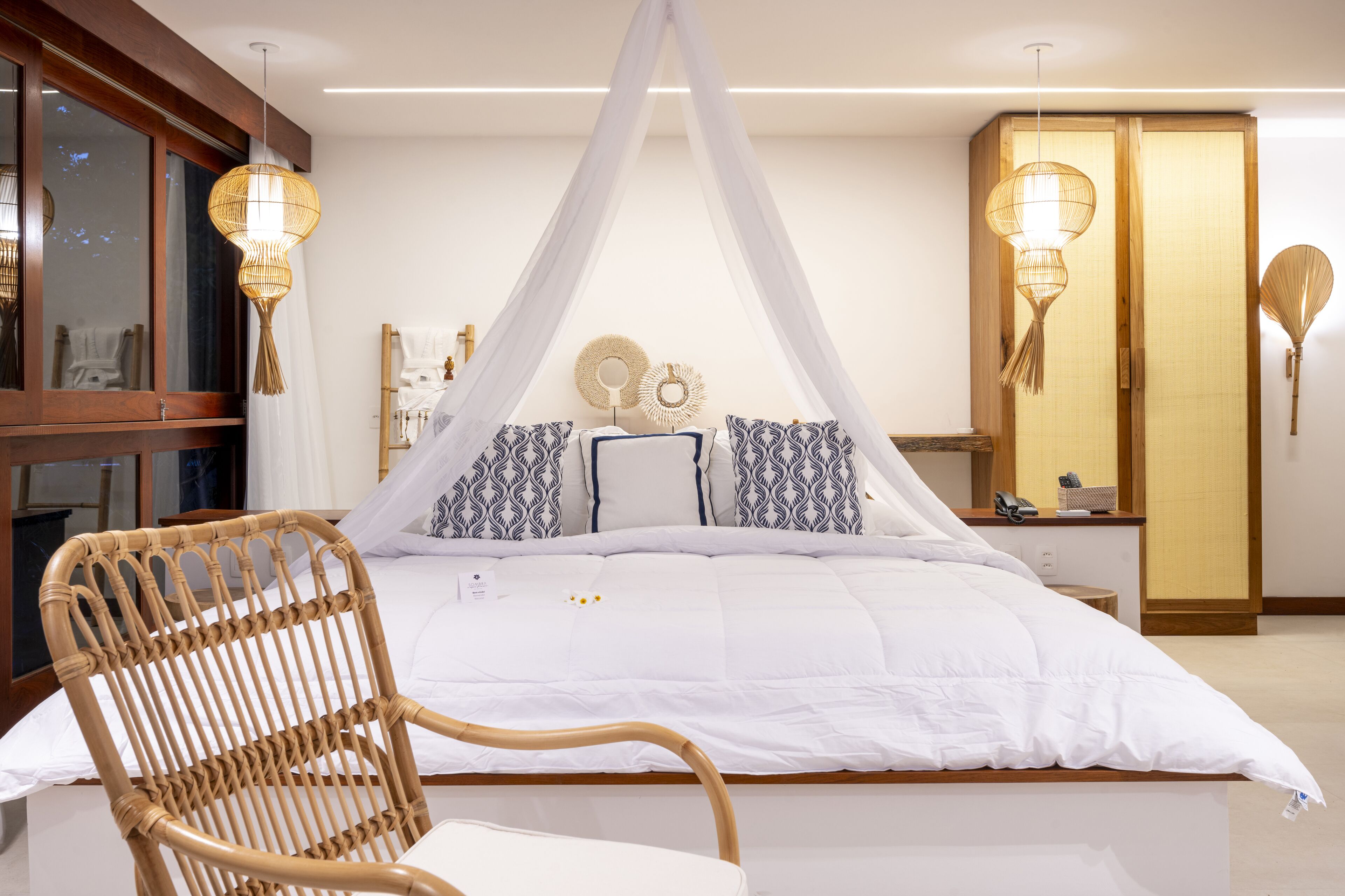 Un dormitorio con cama con dosel, una silla de mimbre, una mesita de noche con lámpara y un armario.
