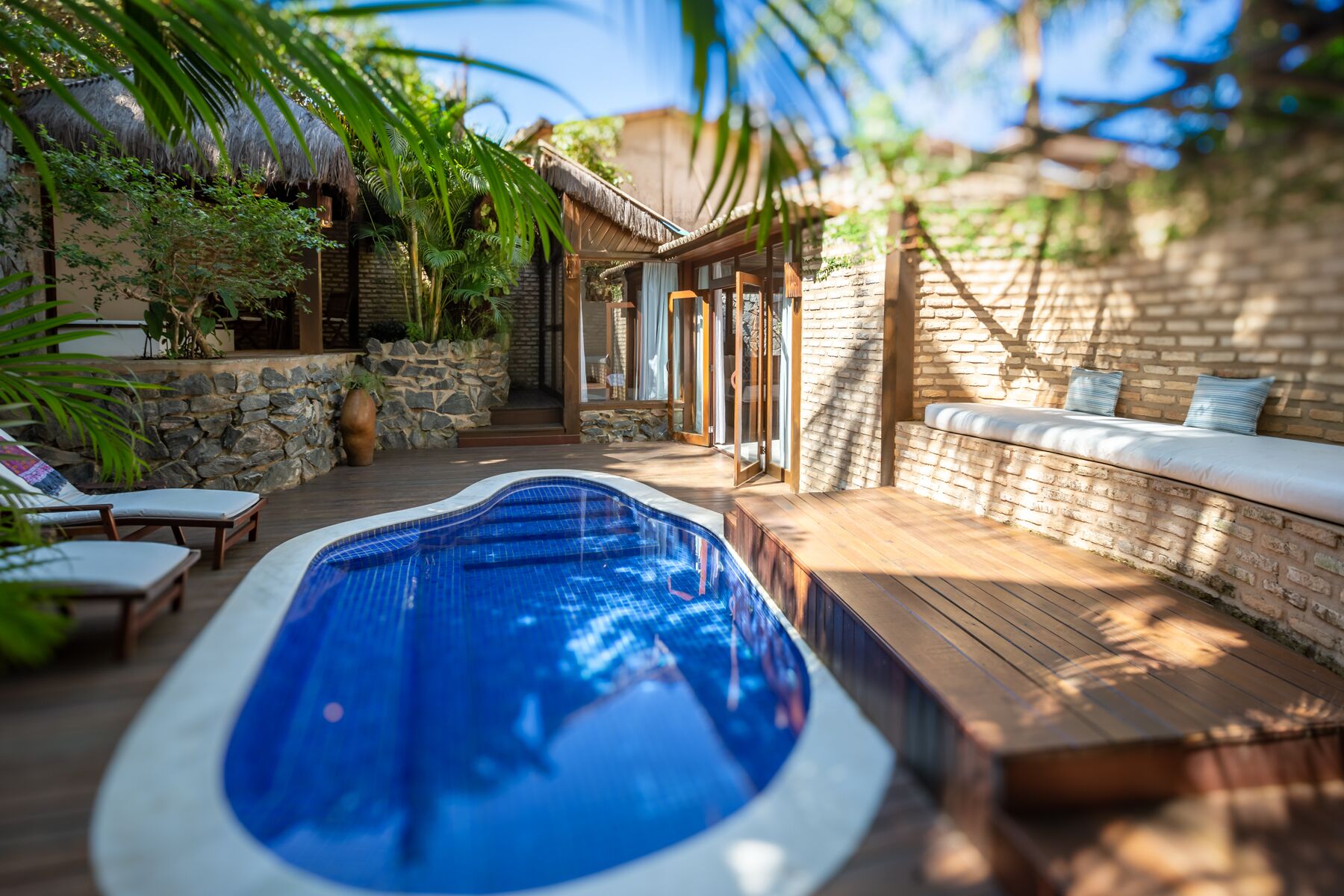 A swimming pool with a wooden deck, stone walls, and a thatched roof structure.