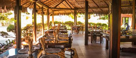 An outdoor dining area with thatched roof, wooden tables, and chairs, surrounded by greenery.